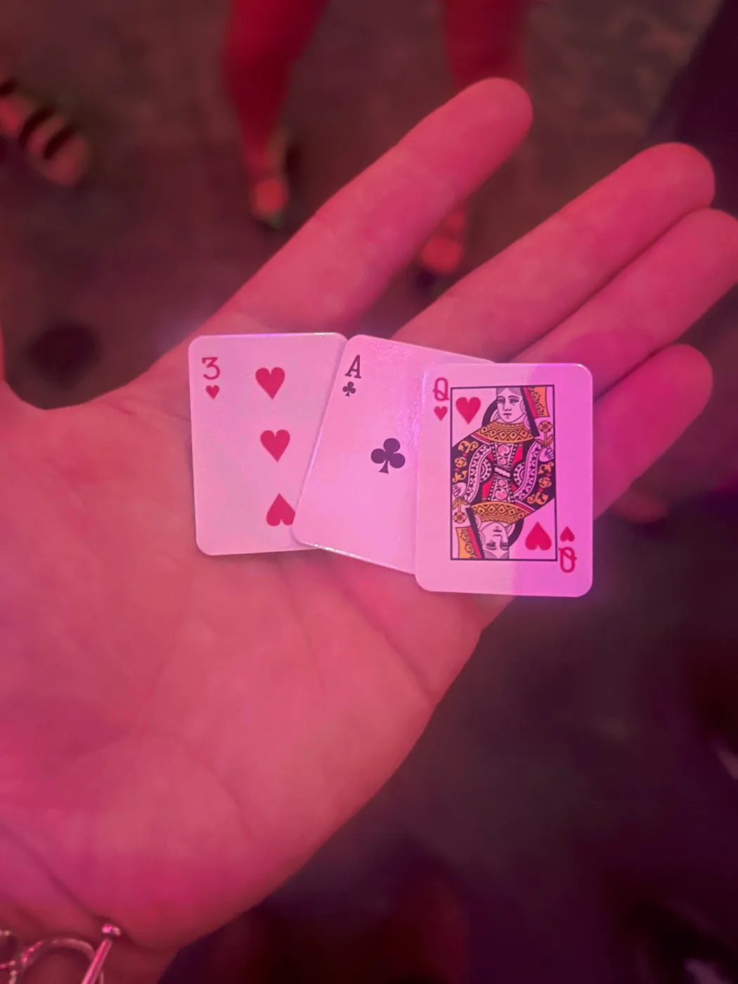 A hand holding three playing cards under pink light: the three of hearts, the ace of clubs and the queen of hearts, reading left to right as 3 A Q.